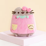 A sitting Pusheen Plush wearing a pink dress with yellow pockets, mint sleeves and a collar, as well as Hello Kitty's bow on top of her head. The Hello Kitty x Pusheen collaboration logo is present on the right dress pocket.