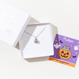 The silver charm necklace in a square white jewelry box. A square card insert is next to the box, featuring artwork of the charm.