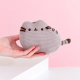 Model holds Knit Pusheen Plush. Mini plush is made with a grey knit fabric thread to give the plush a crocheted look. Grey embroidery is used for Pusheen’s stripes while brown embroidery is used for Pusheen the Cat’s mouth and eyes. Plush measures 6” long and 4” tall.  