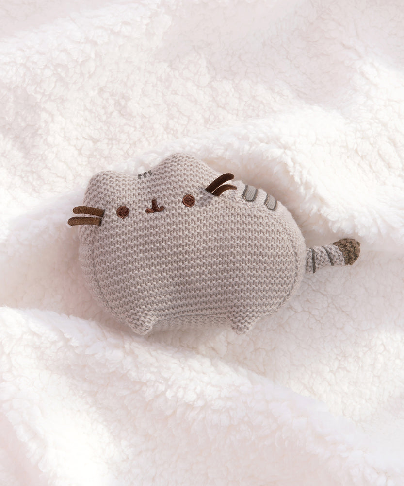 Knit Pusheen Plush lies on a white, fluffy surface. The brown ribbon whiskers extend off the plush face. The plush is sculpted so the cat’s ears and paws are defined. 