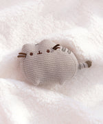 Knit Pusheen Plush lies on a white, fluffy surface. The brown ribbon whiskers extend off the plush face. The plush is sculpted so the cat’s ears and paws are defined. 