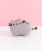 Front view of plush. Knit Pusheen Plush sits atop a white surface in front of a light pink background. The mini plush has a small grey crocheted look to it. 