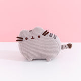 Front view of plush. Knit Pusheen Plush sits atop a white surface in front of a light pink background. The mini plush has a small grey crocheted look to it. 