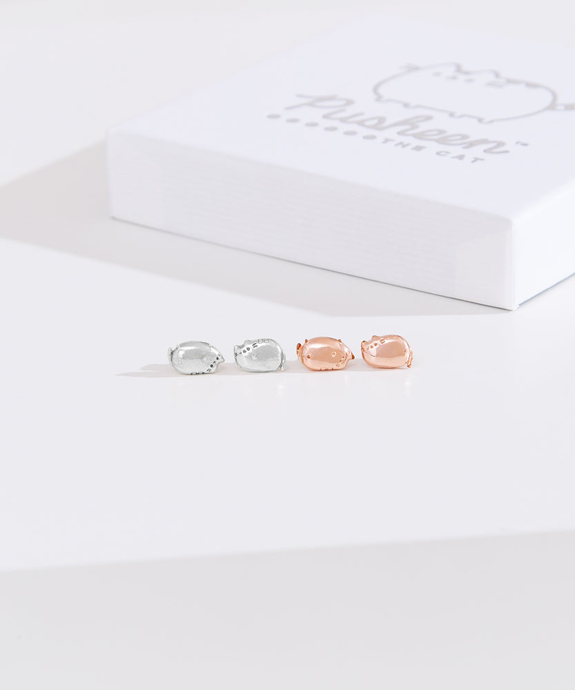 Front view of Pusheen Lazy Stud Earrings, which are unmatched loaf poses of Pusheen the Cat. The earrings on the left are sterling silver and on the right are the rose gold vermeil earrings. Behind the earrings sits the white packaging case that the earrings come in. 