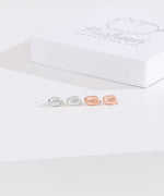 Front view of Pusheen Lazy Stud Earrings, which are unmatched loaf poses of Pusheen the Cat. The earrings on the left are sterling silver and on the right are the rose gold vermeil earrings. Behind the earrings sits the white packaging case that the earrings come in. 
