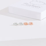 Front view of Pusheen Lazy Stud Earrings, which are unmatched loaf poses of Pusheen the Cat. The earrings on the left are sterling silver and on the right are the rose gold vermeil earrings. Behind the earrings sits the white packaging case that the earrings come in. 