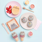 A photo of various Pusheen treats from the cookbook, arranged on plates on top of a light blue surface. Some of the baked goods include Pusheen Pancakes, Pusheen on toast, a dome cookie of Pusheen holding various mini food, and Pusheen peeking out of a sprinkle cake cone.