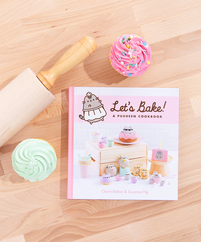 The Pusheen Cookbook laying flat on top of a wooden surface, surrounded by a wooden rolling pin and a pink and mint frosted cupcake. The spine of the book is pink, and by the title of the book is an illustration of Pusheen in a white apron and a chef’s hat.