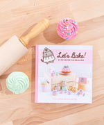 The Pusheen Cookbook laying flat on top of a wooden surface, surrounded by a wooden rolling pin and a pink and mint frosted cupcake. The spine of the book is pink, and by the title of the book is an illustration of Pusheen in a white apron and a chef’s hat.