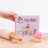 A hand holdind the corner of the Pusheen cook book, standing on top of a wooden surface surrounded by a rolling pin and a pink frosted cupcake.