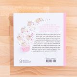View of the back cover of the cook book on top of a wooden surface. The back cover features Pusheen-shaped marshmallows erupting from a pink cupcake cup alongside regular marshmallows and multicolor stars. The back reads ‘A Color and fun collection of PUsheen-themed recipes for cookies, cakes and other delicious treats’ in pink, with more back copy in dark brown. The publisher’s information is noted on the lower left, and the barcode is noted on the lower right.