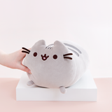 Model’s hand lightly squeezing the Medium Log Squisheen plush, which has been placed on a square white pedestal in front of a light pink and white background. The Medium Log plush features Pusheen laying on her stomach, shaped like a cylinder with two nub paws upfront. Pusheen’s eyes and slightly open mouth are embroidered onto her face, and she has two felt whiskers poking out from each side of her face. 