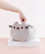 A hand pushing it’s finger straight down onto the back of the Medium Log Squisheen plush, which has been placed on a square white pedestal in front of a light pink and white background. While the back of the plush has been indented, there has not been enough force to distort the face of the plush.
