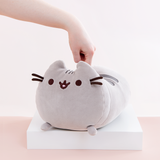 A hand pushing it’s finger straight down onto the back of the Medium Log Squisheen plush, which has been placed on a square white pedestal in front of a light pink and white background. While the back of the plush has been indented, there has not been enough force to distort the face of the plush.