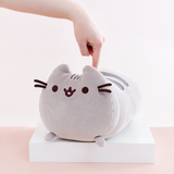 A hand pointing a finger directly on top of the back of the Medium Log Squisheen plush, which has been placed on a square white pedestal in front of a light pink and white background.