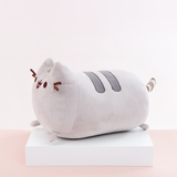 Side view of the Medium Log Squisheen, facing the left, placed on top a square white pedestal in front of a light pink and white background. Pusheen’s back feet and striped tail stick out from her behind.