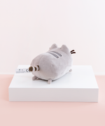 Back quarter view of the Mini Log Squisheen, facing the right, placed on top a square white pedestal in front of a light pink and white background. Pusheen’s behind is just as round as her front. Her striped tail sits directly in the middle of the circle. There are two nub feet at the bottom of the circle, mirroring the ones up front. An information tag sticks out to the left of the tail.