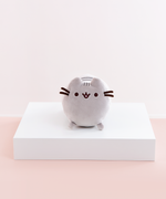 Front view of the Mini Log Squisheen plush, which has been placed on a square white pedestal in front of a light pink and white background. The front of Pusheen is a perfect circle, save for her two ears poking out on top. Pusheen’s face and whiskers are located closer to the top, while her nub feet are at the very bottom. Pusheen’s back stripes are partially visible through the gap between Pusheen’s ears.