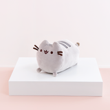 Quarter profile view of the Mini Log Squisheen, facing the left, placed on top a square white pedestal in front of a light pink and white background. Pusheen’s body is shaped like a log, with two dark grey stripes on top of her back, and her back feet and tail partially peeking out from her behind.
