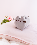 The Mini Log Squisheen rests elegantly on top of a light pink pillow. Pink Peonies and a fuzzy white blanket peek out from the edges of the pillow.