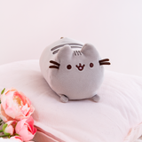 The Mini Log Squisheen takes center stage as she rests elegantly on top of a light pink pillow. Pink Peonies and a fuzzy white blanket peek out from the bottom left.