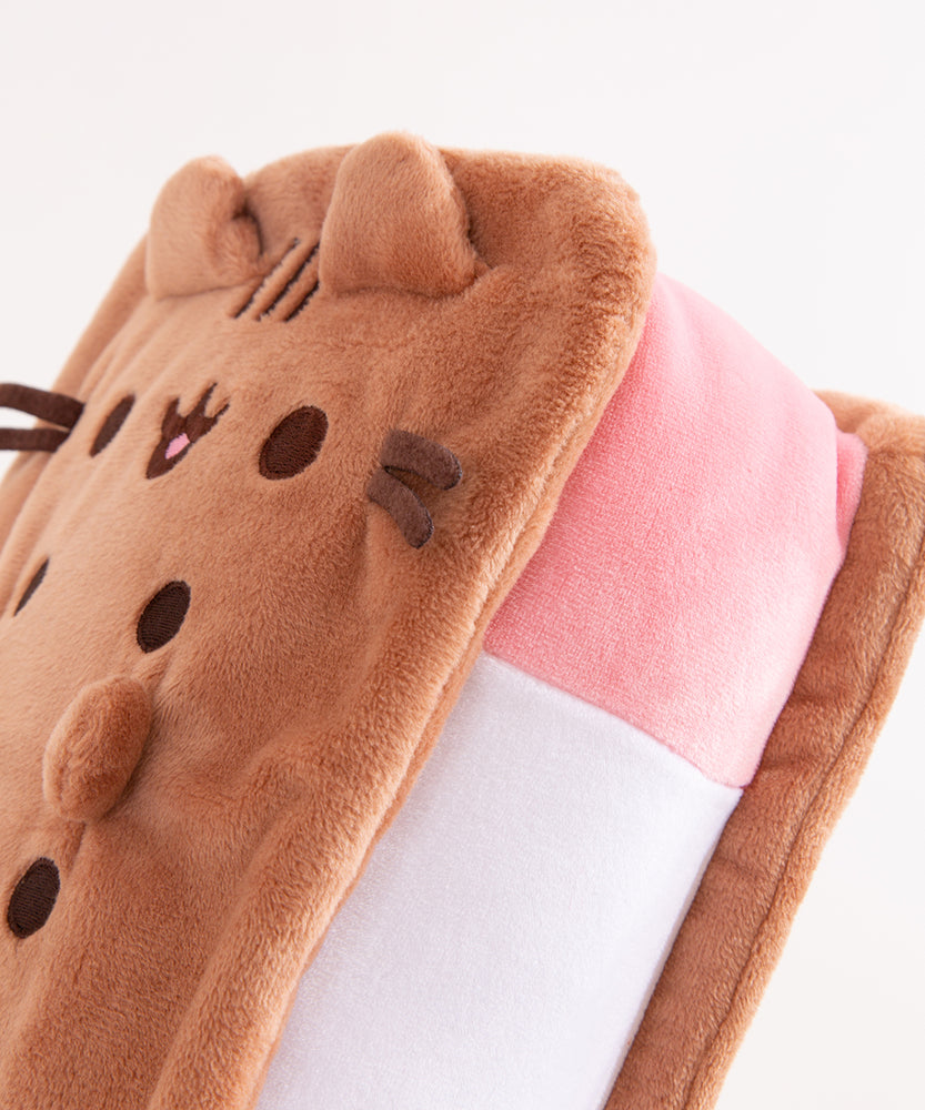 Top view of the squisheen plush shows off Pusheen’s brown face and four paws. Pusheen plush lies on a white surface.  