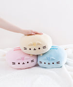 A trio of Round Squisheen plush stacked up in the shape of a triangle, pink and blue at the bottom with the yellow on top. A model’s hand is resting on top of the triangle.