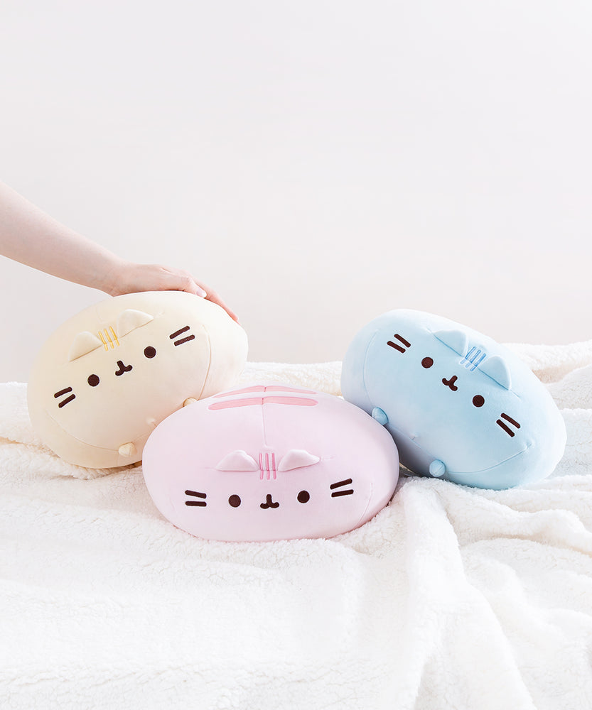 The Round Squisheen Plush trio collapsed on top of a fluffy white blanket, a model’s hand emerging from the left to rest on top of the yellow plush, the pink plush hiding her feet in the middle, and the blue plush on the right nearly on it’s side.