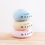 The three Round Squisheen Plush stacked on top of one another, sitting on top a wooden floor in front of a white background. The pink plush is at the bottom, the yellow in the middle, and blue on the top. The pink and yellow plush are squished down enough to look as if they have no feet, but the blue round Pusheen still has her feet.