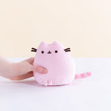 A model’s hand holding the pink Mini Squisheen Plush in front of a white and pale yellow background. The pink plush has dark pink stripes.