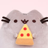 Closeup view of Pizza Pusheen Plush. In her paws, Pusheen holds a yellow, red, and light brown slice of pizza representing pepperoni pizza. She is shown with her classic black and brown nose, eyes, and whiskers.