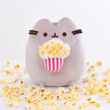 Pusheen sitting upright, her two top nub paws holding a plush striped bag of popcorn, and her bottom nub feet rest at the bottom. The popcorn in the plush basket is made of a different material that resembles cotton balls, and has butter slathered all over it. The plush is surrounded by loose pieces of popcorn.