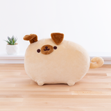 The Pugsheen plush in front of a white wall on top of a wooden floor, and a potted succulent can be seen to the plush’s left. Pugsheen’s left ear is slightly raised, as if something has caught her attention. The inside of her ear is the same brown fabric as the rest of the ear and her snout.