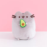 Front view of Pusheen Avocado Plush. Pusheen the Cat is shown with her classic brown eyes, mouth, and whiskers. Pusheen holds a cut-in-half, two-toned green avocado with a light brown pit.