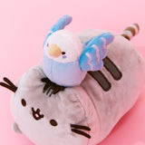 Quarter view closeup of the Bo and Pusheen plush. Bo's eyes, cheeks and features in her wings are embroidered. Bo has a round head, and the white coloring goes down to the top of her wings. Pusheen's face and stripes are embroidered, and her whiskers are two felt stripes on each cheek.