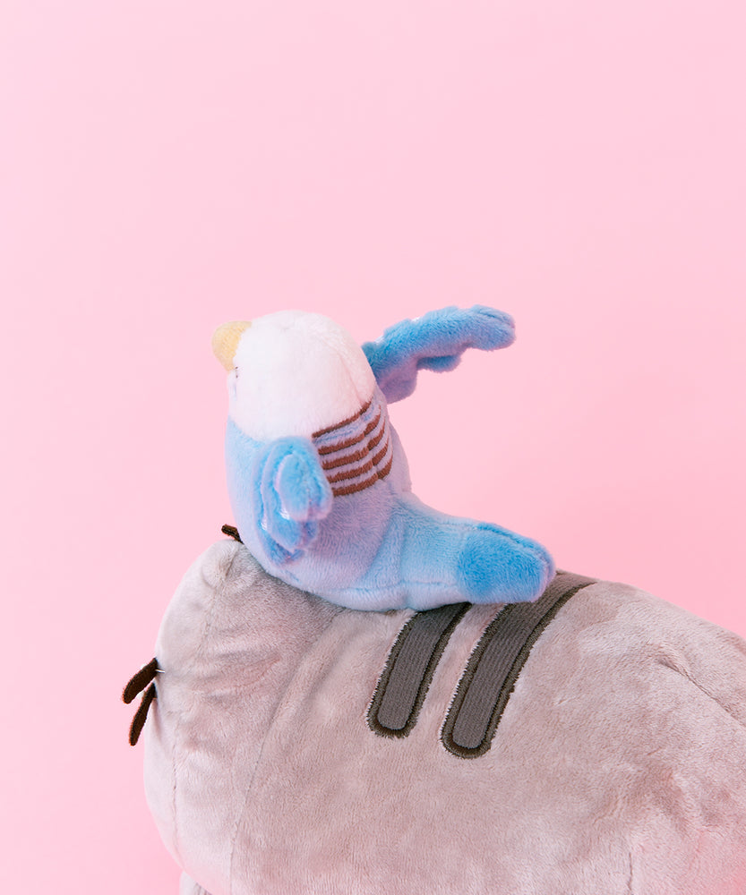 Back Quarter view close up of the Bo and Pusheen plush. Bo's back features five light grey stripes on her back, and a light blue tail that reaches out to the second stripe on Pusheen's back. Bo's tail is rounded off.