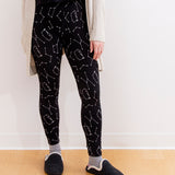 Model standing while wearing the Pusheen Celestial Leggings. The model is wearing a loose knit sweater and black slippers with a white trim. The model is standing in front of a white wall and a wooden floor.