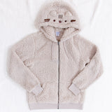 A fuzzy light grey zip up hoodie with cat ears and Pusheen's face on the hood. There are drawstrings on the hoodie, pockets, and the cuff and waistband are ribbed fabric. The hoodie is laying on top of a fuzzy white blanket with the sleeves folded inwards.