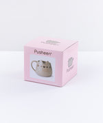 The box packaging for the Pusheen Character Mug in front of a white background. The box is a pink cardboard square that cover the mug entirely. On the front of the box is the Pusheen logo, a photo of the mug inside, and the name ‘Ceramic Mug’ printed underneath it in English and French. The top and side of the box features the Pusheen the Cat Logo.