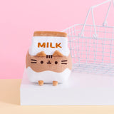 The Pusheen Chocolate Milk Sips Plush seated on a white pedestal with a small white wire basket in the background. The plush is in the shape of a small milk carton with a brown and white wave throughout the body and the word ‘Milk’ embroidered in brown stitching near the carton lid. Pusheen’s face is embroidered in dark brown on the plush, with plush ears at the rim of the carton lid and brown plush feet at the bottom. 