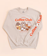   Front and sleeve view of the tan unisex sweatshirt. The arms are folded in to the center of the sweatshirt to reveal the printed sleeve detail. The sleeve features the phrase “Coffee O’ Clock” with Pusheen sitting in a white coffee cup surrounded by two coffee beans.   