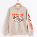 Front view of the Coffee Club Unisex Sweatshirt. The sand/tan sweatshirt hangs on a brown wood hanger to show all collar, sleeve, and hem details. 
