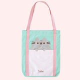 A tote bag is propped up on a pink surface. The mint green tote has an applique of Pusheen the Cat with her paws hanging over the front pocket. The handles of the bag are pink, and the base has the Pusheen the Cat logo in brown. 