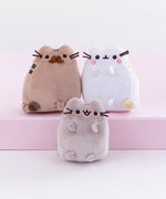 Close up of the mini plush, Pusheen’s parents on top of the pink pedestal while Pusheen sits underneath them on the white floor. The parent’s poses mirror each other, with the father’s tail pointing out from the left and the mother’s tail pointing out from the right. Pusheen faces straight forward, no tail visible and mouth open.