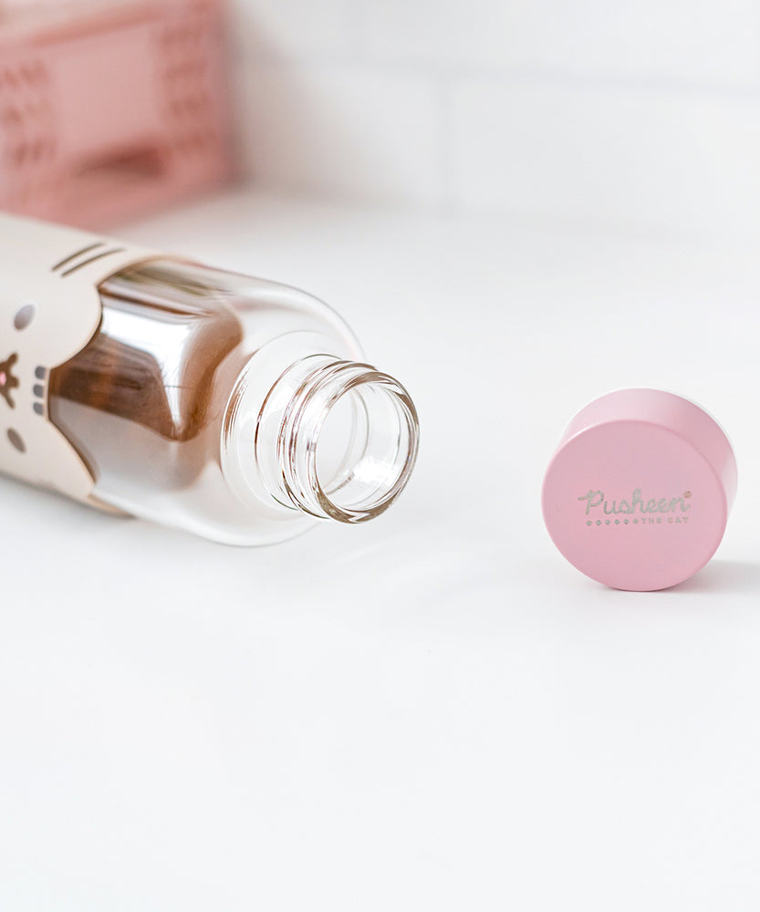 Glass water bottle lies on a white surface showing the inside of the open bottle. The pink top features a Pusheen the Cat logo in silver print.  