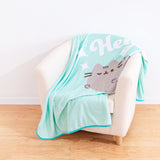 Pusheen Hey Throw Blanket draped on top of a cream chair. The teal blanket features a graphic of Pusheen the Cat winking while saying “Hey.” The center graphic is surrounded by white stars.  