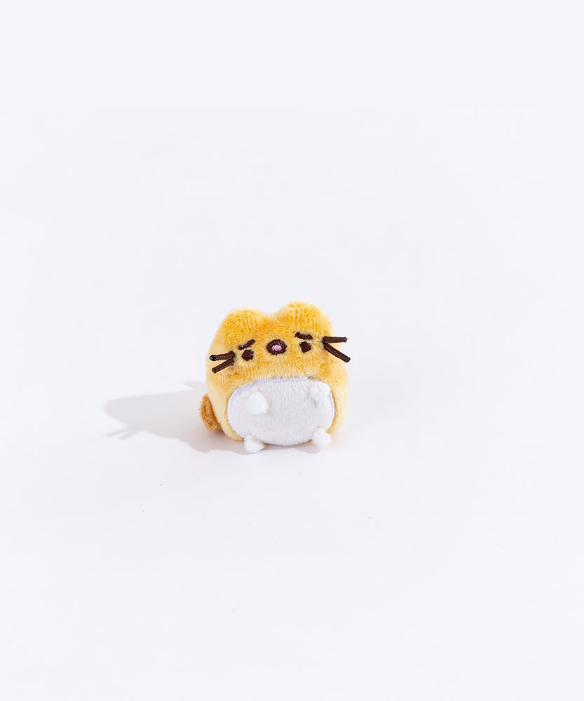 Full view of the mini worried yellow cat plush from the collectors set. The cat is a golden yellow, with a dark orange tail on the left side. Their belly has a white circle, which encompasses their feet. The cat has worried eyebrows embroidered into it, and their mouth is open.