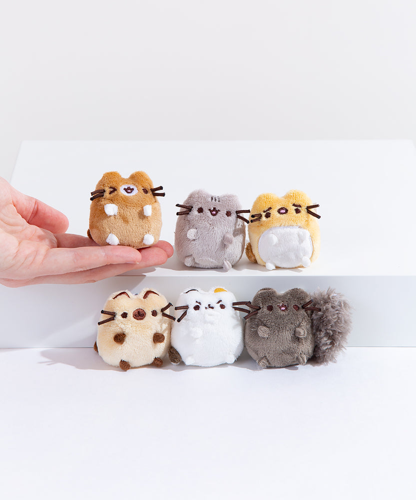 The I Love Kitties mini plush lined up in two rows of 3, a model’s hand holding the winking brown mini cat plush on the tip of their fingers. All of the mini plush have all of their paws sticking out forward, save for the annoyed white cat who has one paw over their mouth and nose.