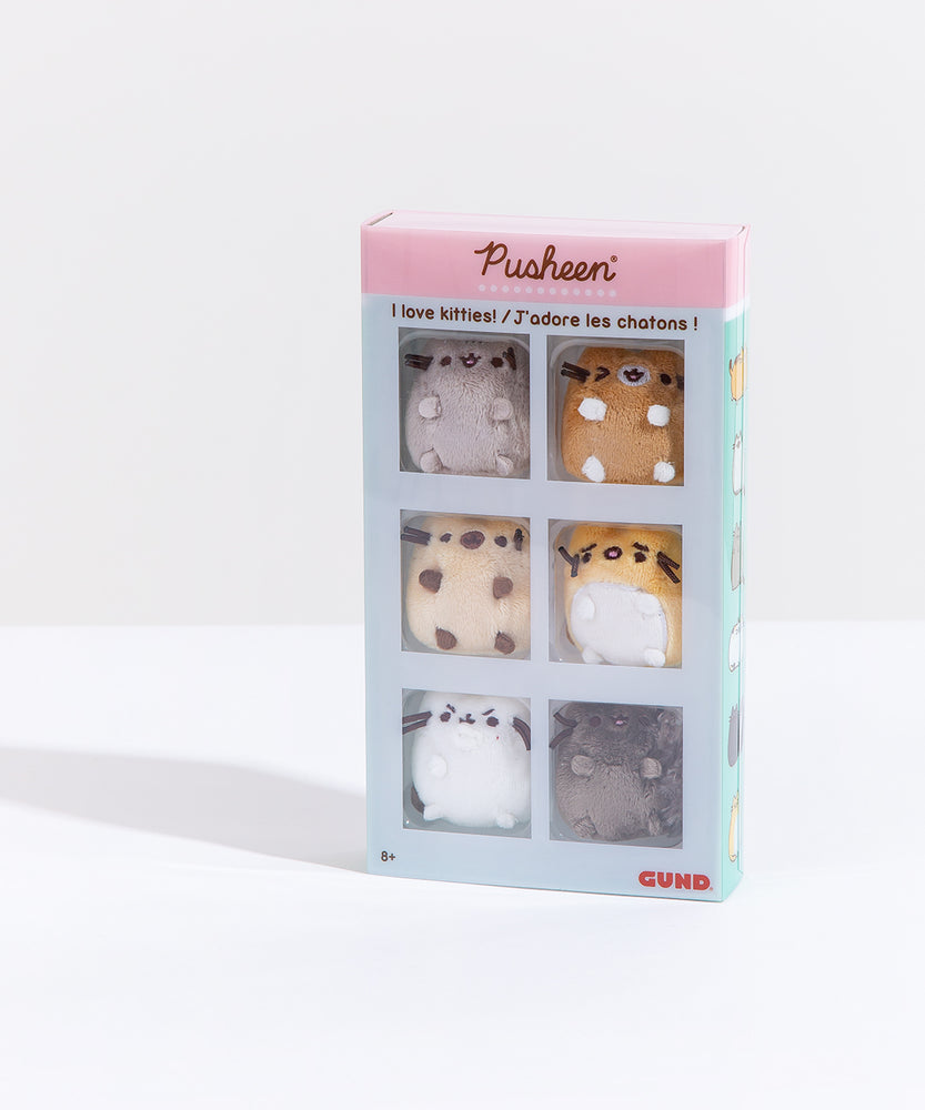 The Pusheen I Love Kitties Plush Collectors Set in it’s original packaging standing upright in front of a white background. The plastic sleeve on top of the set features the Pusheen logo on the top pink banner, the text ‘I love kitties!’ in English and French underneath it. The white bars on the sleeve separate the mini plush into their own square, making the set look like a comic strip. The 8+ age rating is noted on the bottom left, and the GUND manufacturer logo is on the bottom right.