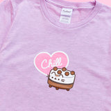 A close-up view of the tee’s screen print graphic. Ice Cream Cake Pusheen is a short cylinder shape with white and brown striped details with light brown balls atop the white top. The ice cream cake features Pusheens’ face printed on the front, her striped tail sticking out from the back, her paws at the bottom of the cake. The word ‘Chill’ is printed in white in a light pink heart with a white outline. The pink heart accompanies the ice cream cake in the center of the tee shirt. 